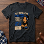 A black graphic t-shirt laid flat on a dark wood background, featuring a printed design with the text 'SOY CATRACHA NO DISCUTO'.