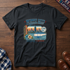 A black short-sleeve graphic t-shirt laid flat on a dark wood background, featuring a colorful vintage-style Chicago skyline and 'Windy City Catracho Heart' print.