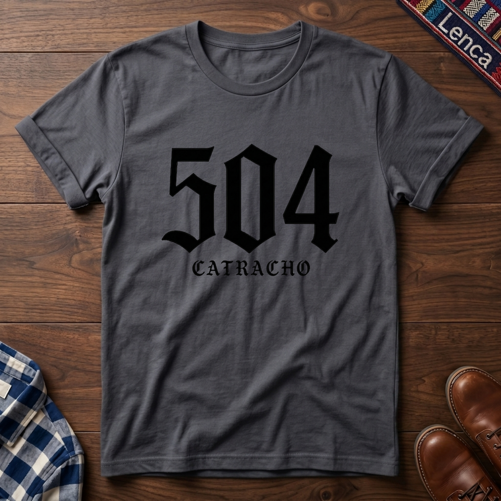A charcoal crewneck t-shirt with a black graphic print reading '504 CATRACHO' in a gothic font, laid flat on a wooden surface.