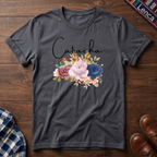 Charcoal short-sleeve crewneck t-shirt laid flat on a dark wood background, featuring the name 'Catacha' in black script above a watercolor floral design with pink, blue, and peach flowers.
