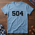A light blue crewneck t-shirt with a black graphic print reading 'HONDURAS 504' and five stars, laid flat on a dark wood background.