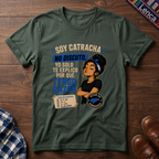 A military green graphic t-shirt laid flat on a dark wood background, featuring a printed design with the text 'SOY CATRACHA NO DISCUTO'.