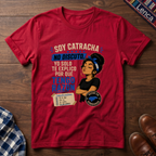 A red graphic t-shirt laid flat on a dark wood background, featuring a printed design with the text 'SOY CATRACHA NO DISCUTO'.