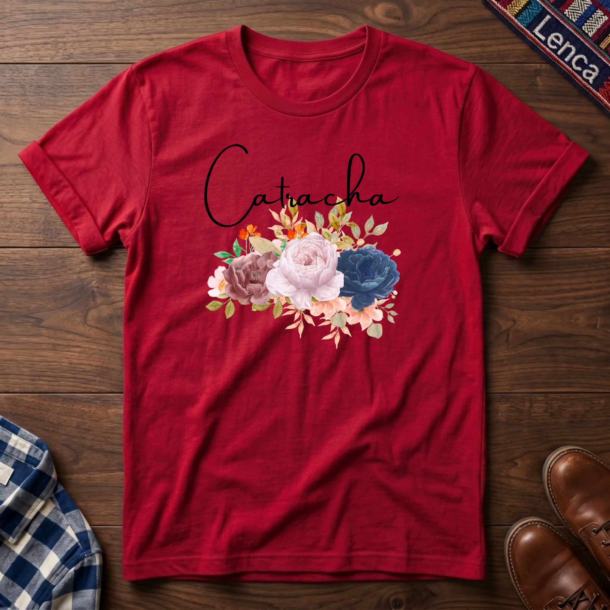 Red short-sleeve crewneck t-shirt laid flat on a dark wood background, featuring the name 'Catacha' in black script above a watercolor floral design with pink, blue, and peach flowers.