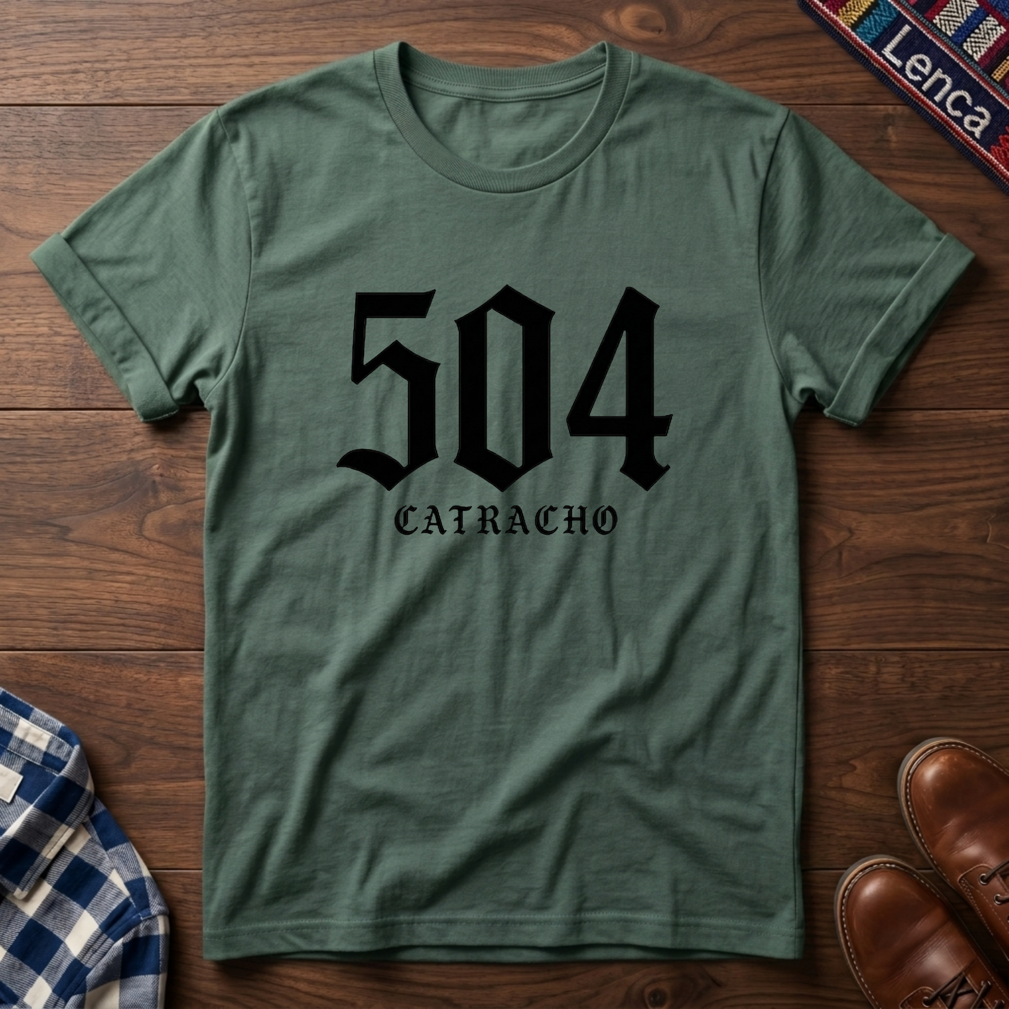 Military green t-shirt with '504 Catracho' text on a wooden surface.