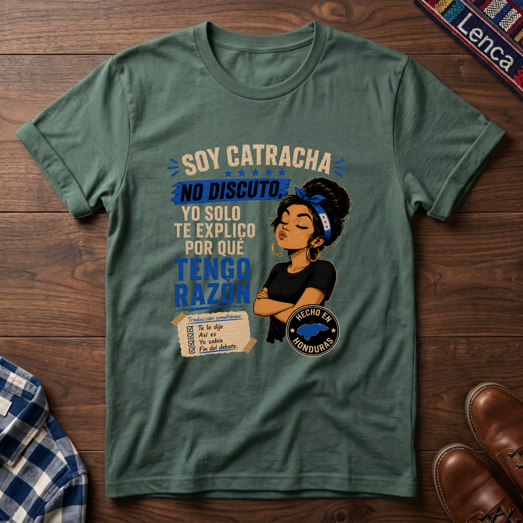 A sage graphic t-shirt laid flat on a dark wood background, featuring a printed design with the text 'SOY CATRACHA NO DISCUTO'.