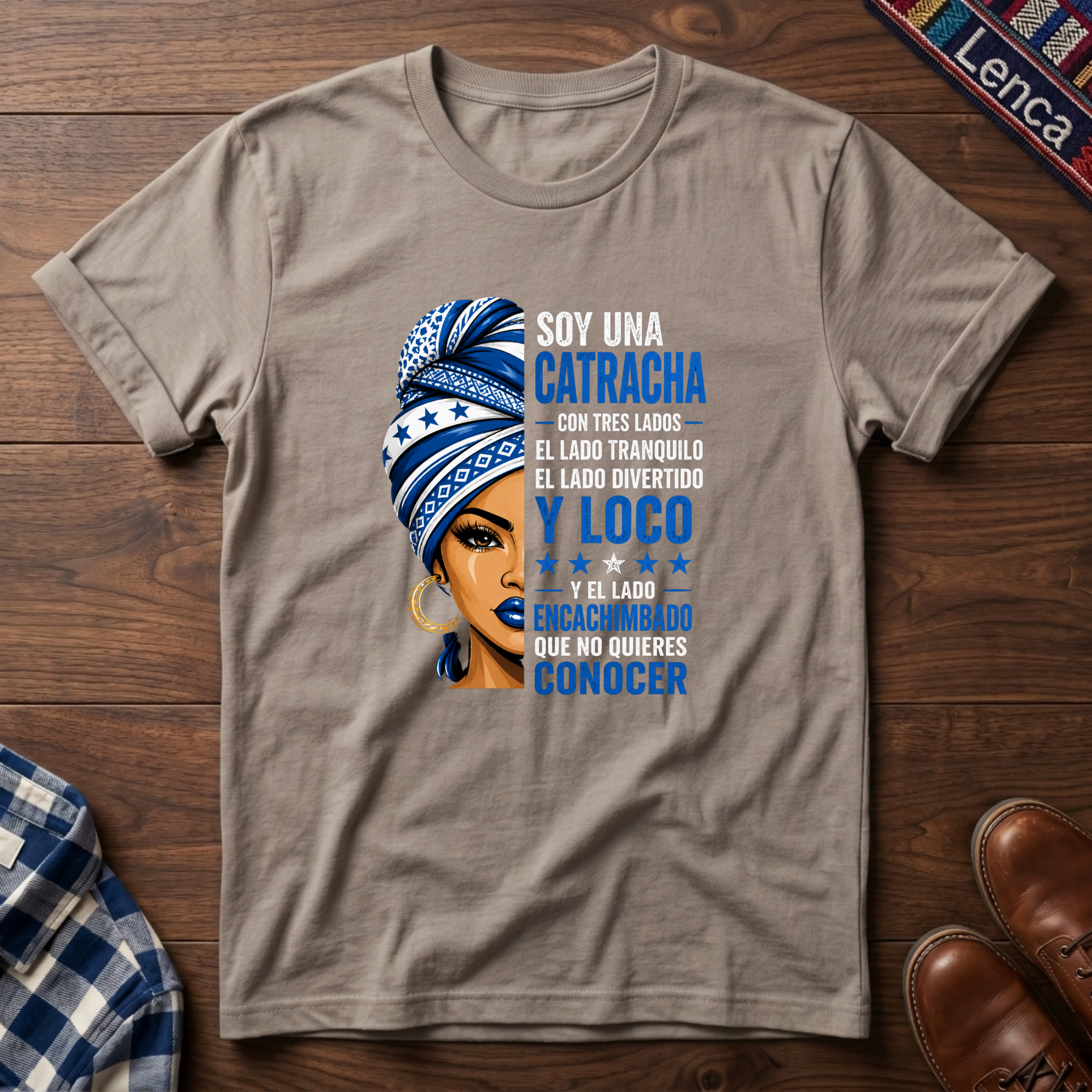 Sand t-shirt with a graphic of a woman with a headwrap and 'Soy Una Catracha' text on a wooden surface.