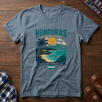 A slate blue graphic t-shirt laid flat on a dark wood background, featuring a colorful tropical Honduras illustration with palm trees, a beach, and mountains. 