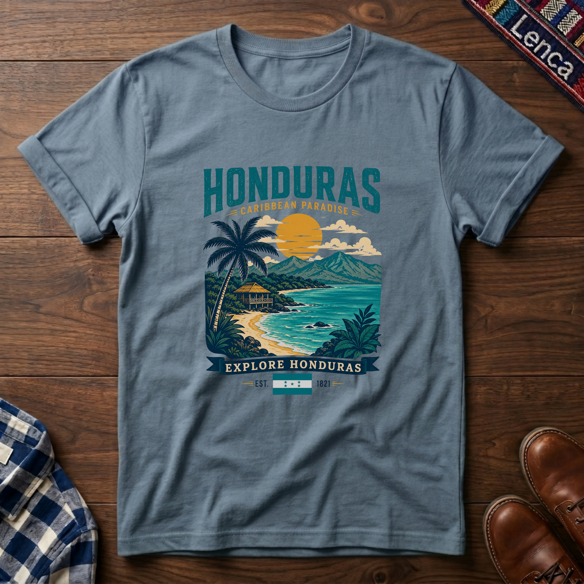 A slate blue graphic t-shirt laid flat on a dark wood background, featuring a colorful tropical Honduras illustration with palm trees, a beach, and mountains. 