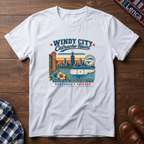 A white short-sleeve graphic t-shirt laid flat on a dark wood background, featuring a colorful vintage-style Chicago skyline and 'Windy City Catracho Heart' print.